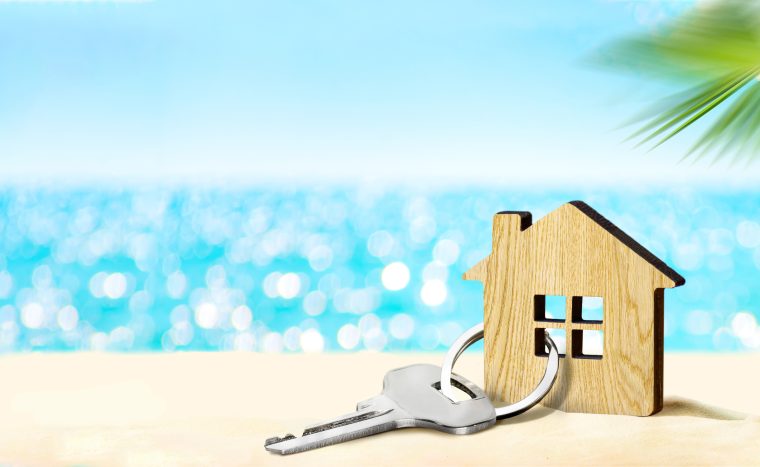 In the foreground, a key attached to a wooden house rests in the sand. The background shows a shimmering blue ocean and part of a palm tree.