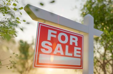 Close up of a red "For Sale" sign with white lettering against a background of trees and sunshine. Stock photo for blog on reasons your house won't sell.
