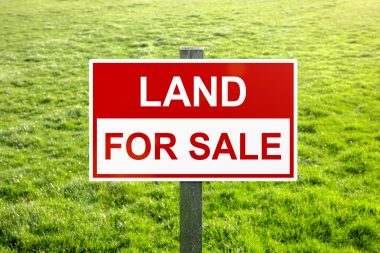 A red and white sign that reads "LAND FOR SALE" in front of a grassy space. 