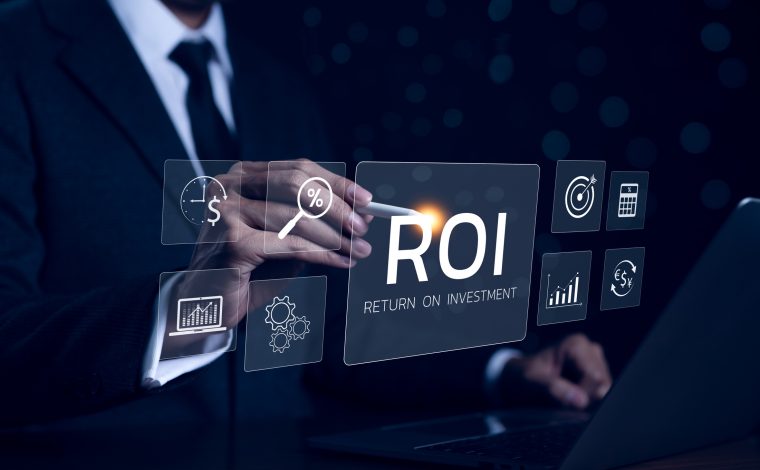 A person in a dark suit and tie in the background points with a stylus to a glowing rectangle that reads "ROI: Return on investment" in the foreground. Other smaller squares contain a target, clock, gears, graph and other icons symbolizing investment.