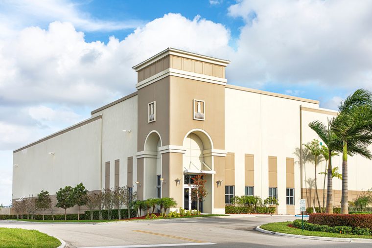 Large beige industrial warehouse with an arched entrance, surrounded by landscaped greenery and palm trees under a bright blue sky with scattered clouds.