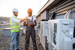 Two workers are on a rooftop inspecting air conditioning units. One is wearing a yellow vest and white helmet, gesturing, while the other holds a clipboard.